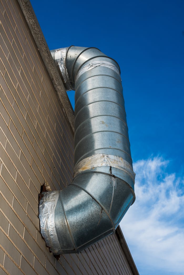 A large industrial air duct attached to a brick wall under a bright blue sky.